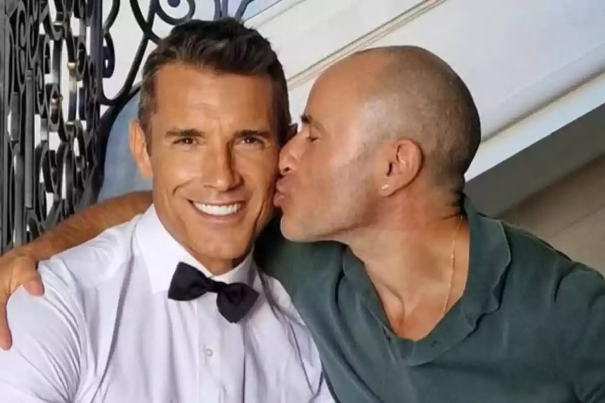 Dos hombres posan juntos, uno de ellos sonríe a la cámara mientras el otro le da un beso en la mejilla Dos hombres posan juntos, uno de ellos sonríe a la cámara mientras el otro le da un beso en la mejilla