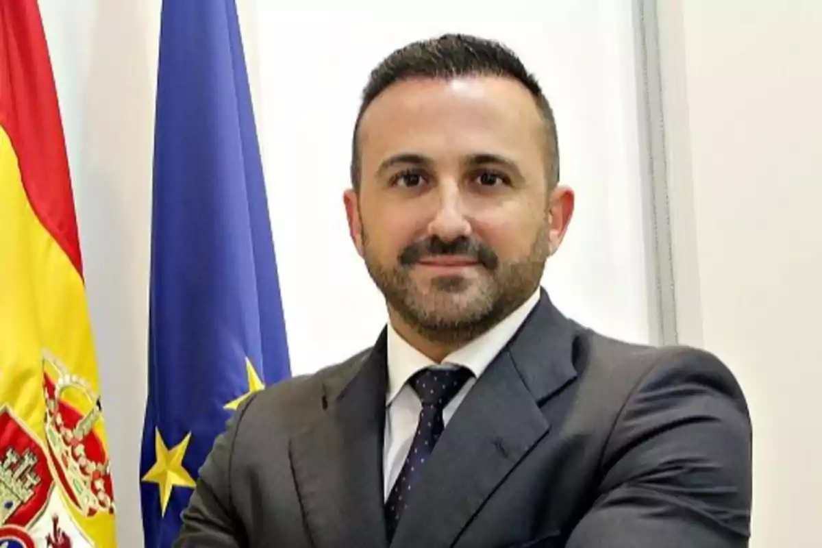 Un hombre con traje y corbata posando frente a las banderas de España y la Unión Europea.