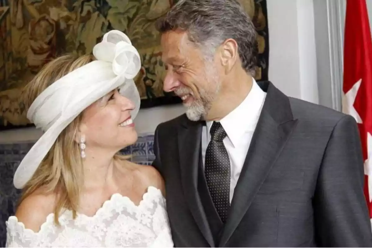 Una pareja sonriente se mira con cariño, ella lleva un vestido blanco con encaje y un sombrero elegante, mientras él viste un traje oscuro con corbata.