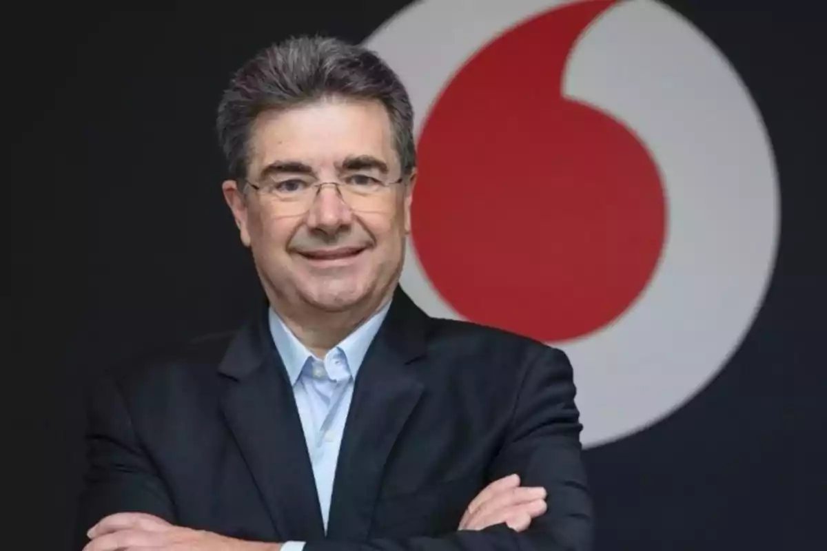 Un hombre con gafas y traje oscuro sonríe frente a un logotipo circular rojo y blanco.