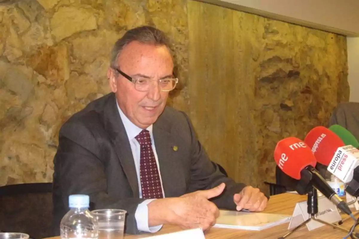 Hombre mayor con gafas y traje oscuro sentado en una mesa frente a varios micrófonos de prensa y una botella de agua