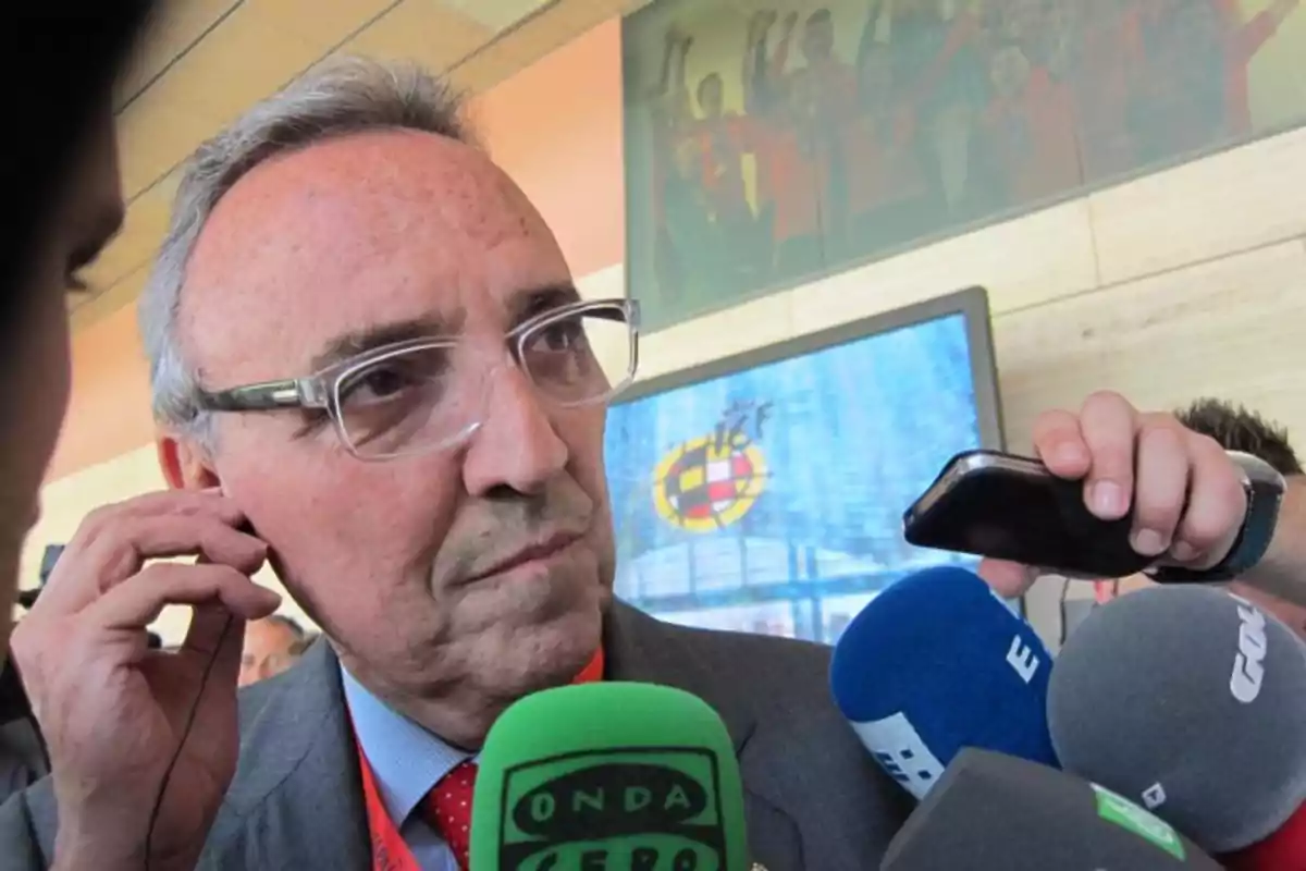 Hombre mayor con gafas siendo entrevistado por varios medios de comunicación frente a un monitor y un cartel deportivo Hombre mayor con gafas siendo entrevistado por varios medios de comunicación frente a un monitor y un cartel deportivo