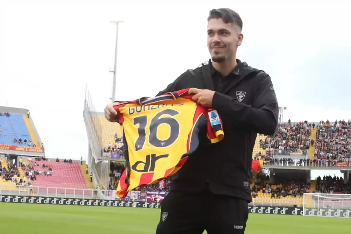 Un futbolista sonriente sostiene una camiseta amarilla y roja con el número 16 en un estadio lleno de aficionados Un futbolista sonriente sostiene una camiseta amarilla y roja con el número 16 en un estadio lleno de aficionados