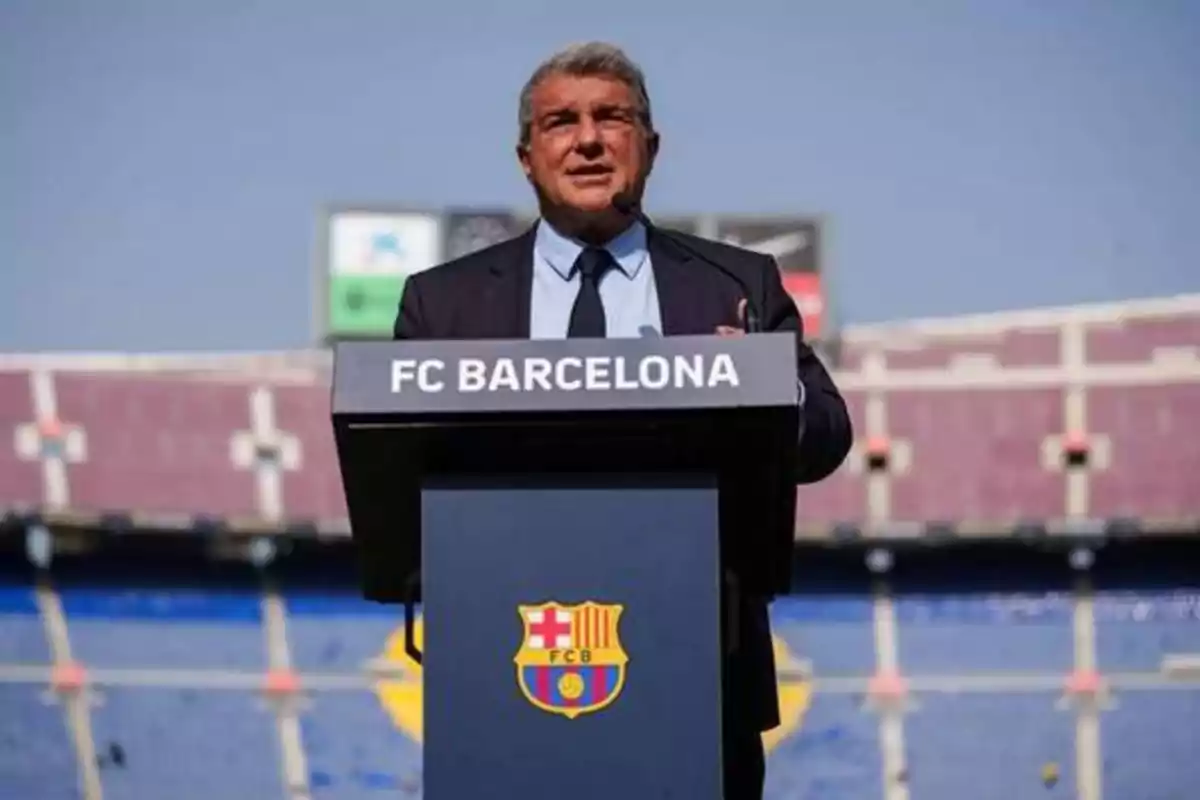 Hombre de traje dando un discurso en un podio con el logo del FC Barcelona en un estadio.