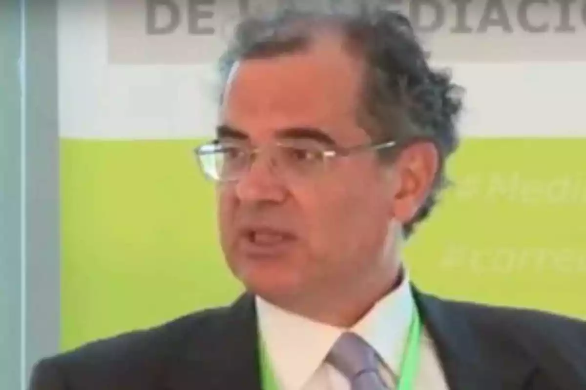 Hombre con gafas y traje hablando en un evento con fondo verde.