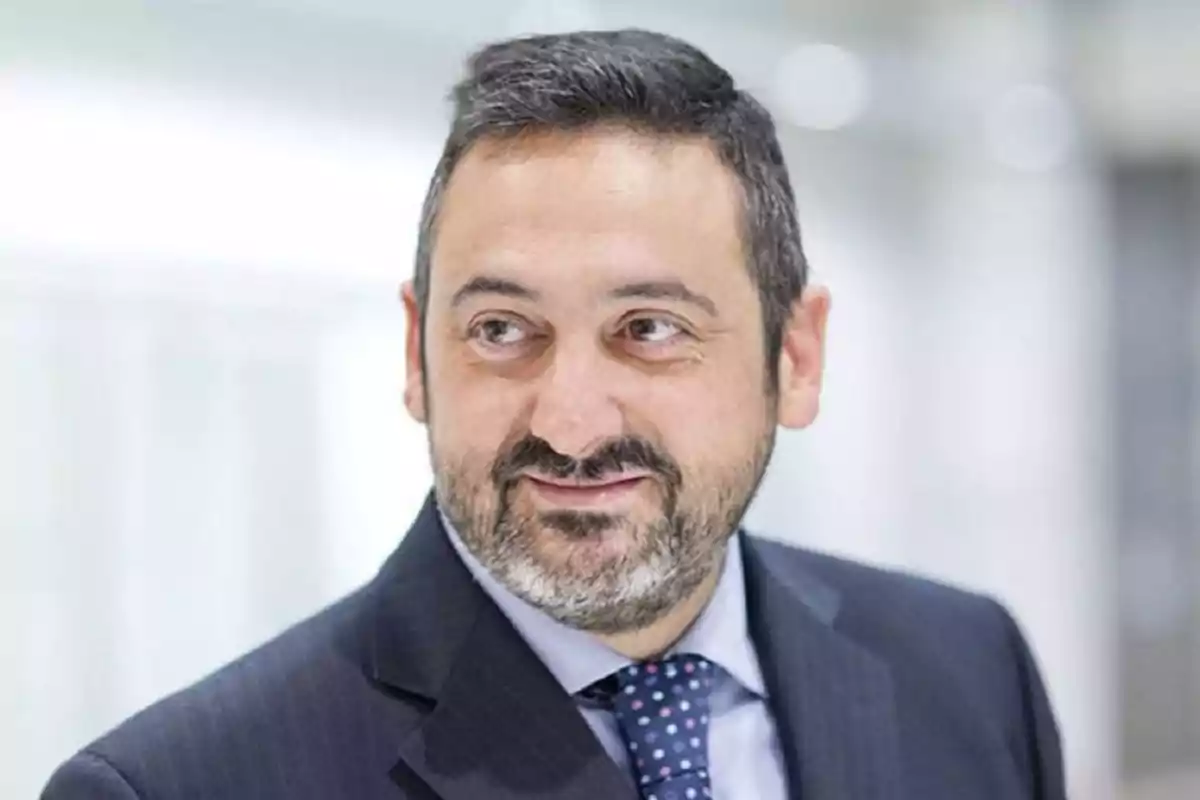 Un hombre con barba y traje oscuro sonriendo ligeramente en un entorno profesional.