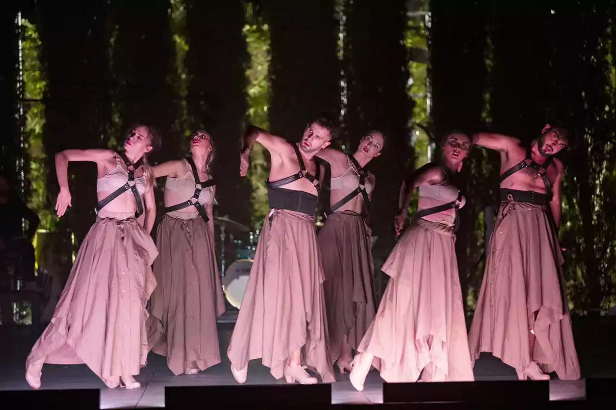 Un grupo de bailarines en el escenario realizando una coreografía con vestuario de tonos neutros y una iluminación tenue.