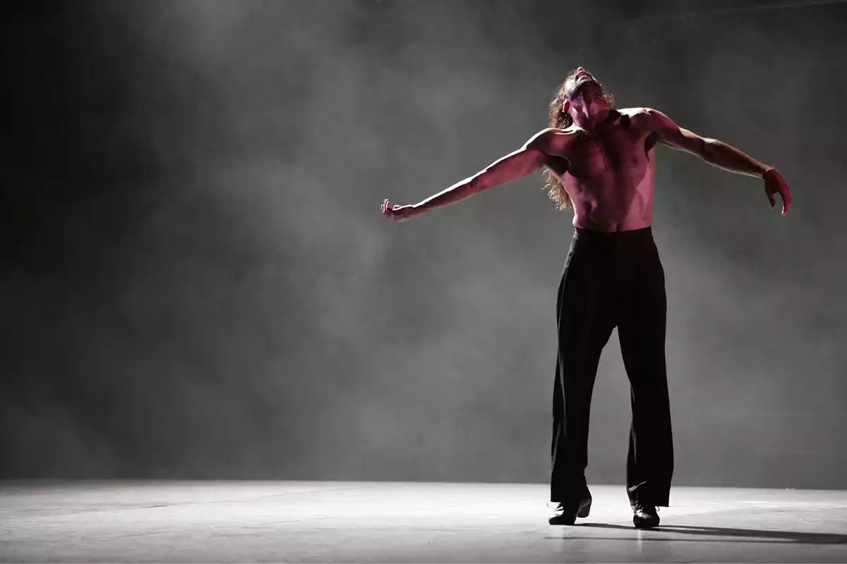 Un bailarín con el torso desnudo y pantalones oscuros realiza una pose dramática en un escenario con humo de fondo.