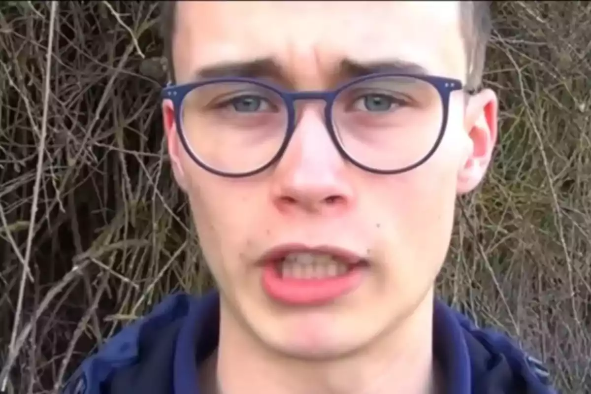 Un joven con gafas de montura azul frente a un fondo de ramas.