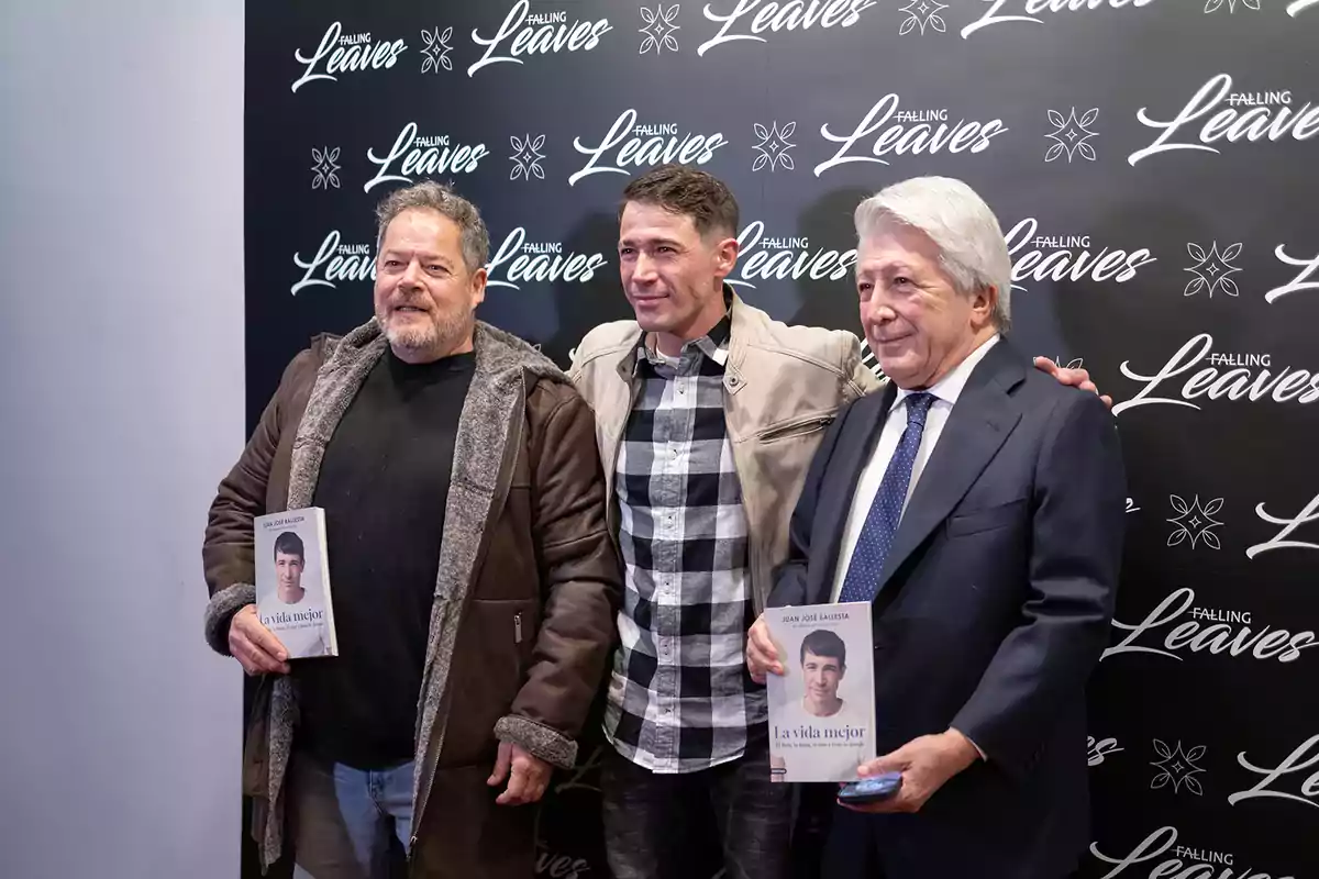 Tres hombres posan juntos en un evento, dos de ellos sostienen un libro titulado 