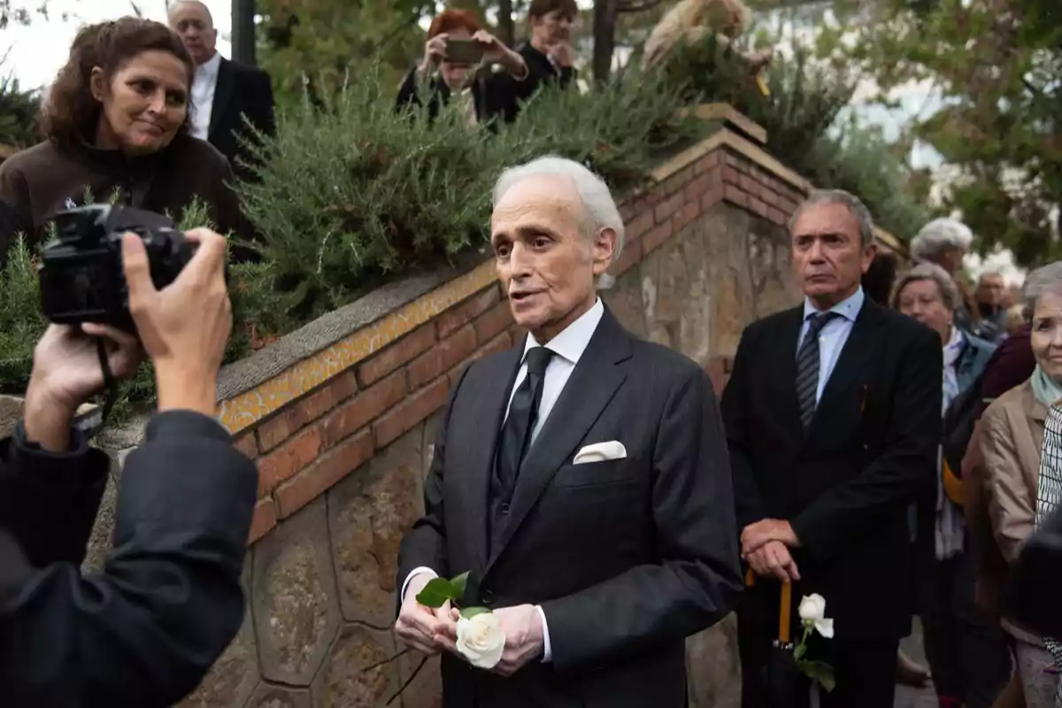 Un hombre mayor vestido de traje oscuro sostiene una rosa blanca mientras es fotografiado en un evento al aire libre, rodeado de otras personas. Un hombre mayor vestido de traje oscuro sostiene una rosa blanca mientras es fotografiado en un evento al aire libre, rodeado de otras personas.
