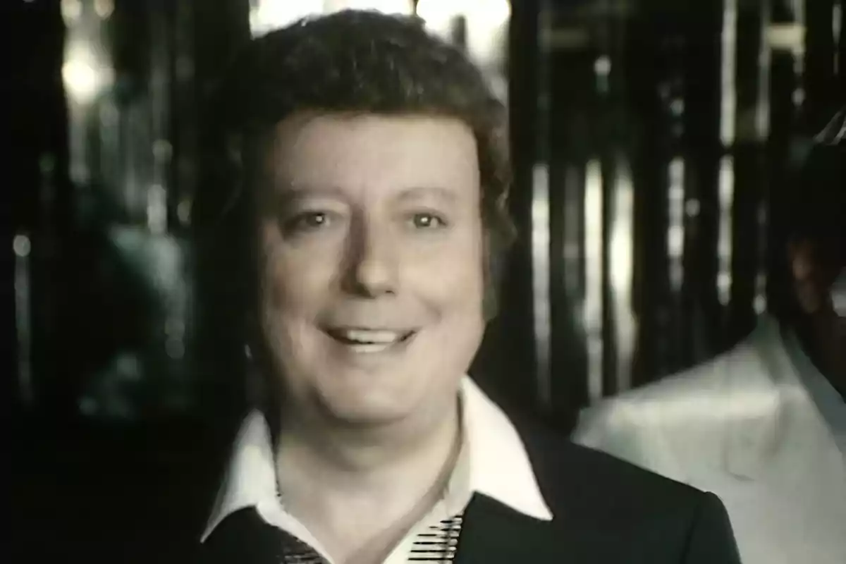 Hombre de cabello rizado y corto sonríe mientras viste una camisa blanca con cuello ancho y un saco oscuro en un ambiente con luces tenues y reflejos metálicos al fondo