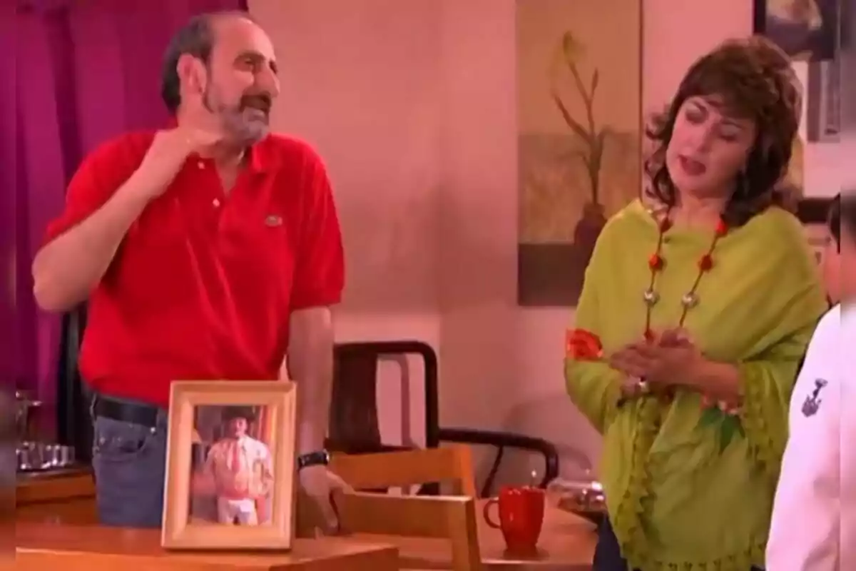 José Luis Gil interpretando a Juan Cuesta con camisa roja y una mujer con blusa verde están conversando en una habitación, sobre una mesa hay un portarretratos y una taza roja. José Luis Gil interpretando a Juan Cuesta con camisa roja y una mujer con blusa verde están conversando en una habitación, sobre una mesa hay un portarretratos y una taza roja.