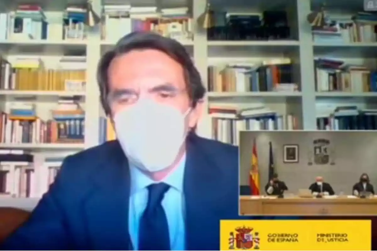 Un hombre con mascarilla blanca y traje oscuro aparece en una videollamada, con una estantería llena de libros detrás de él; en la esquina inferior derecha se ve una pantalla pequeña con una reunión del Ministerio de Justicia del Gobierno de España.