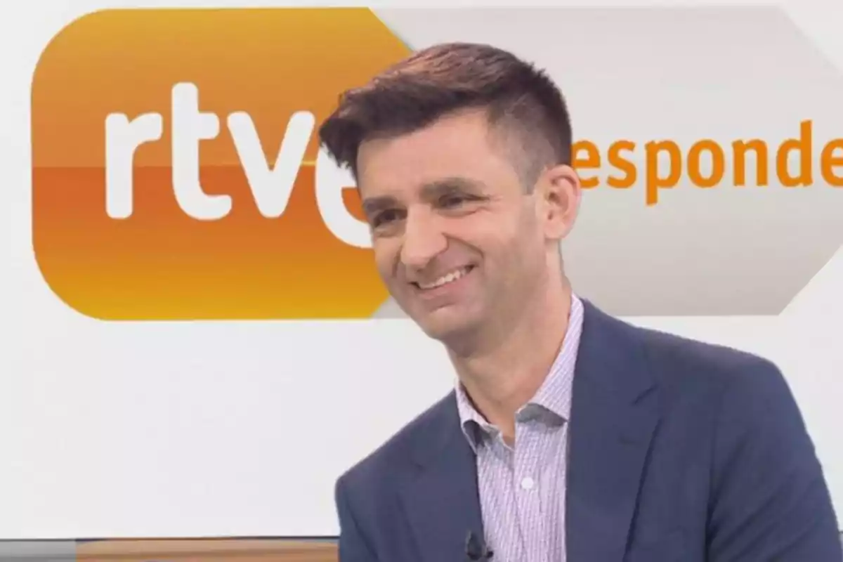 Un hombre sonriente con traje y camisa frente a un fondo con el logotipo de RTVE.