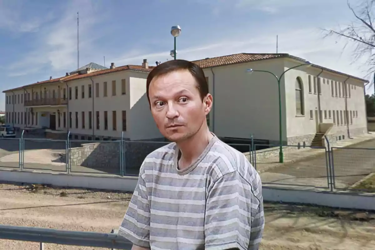 Un hombre con camiseta de rayas grises y blancas está de pie frente a un edificio grande de color claro con techo de tejas rojas y varias ventanas.