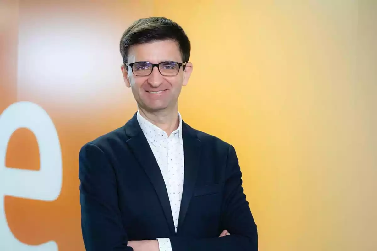 Un hombre con gafas y traje oscuro sonríe frente a un fondo amarillo. Un hombre con gafas y traje oscuro sonríe frente a un fondo amarillo.