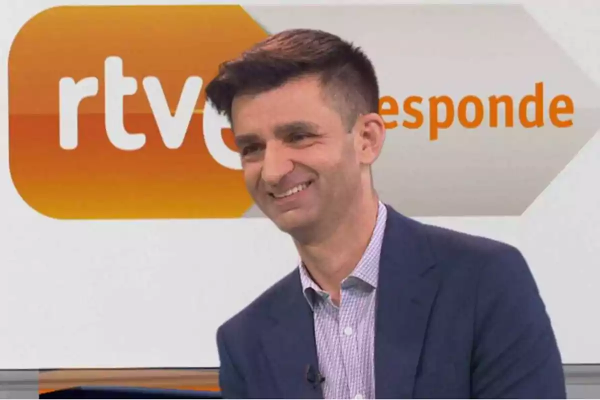 Un hombre sonriente con traje y camisa a cuadros frente a un fondo con el logotipo de RTVE y la palabra 