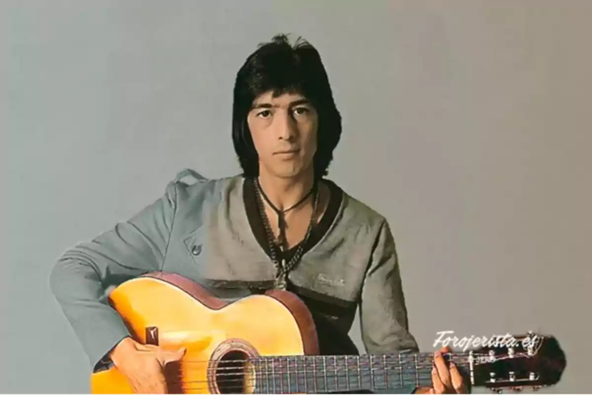 Un hombre con cabello oscuro sostiene una guitarra acústica mientras mira hacia la cámara.