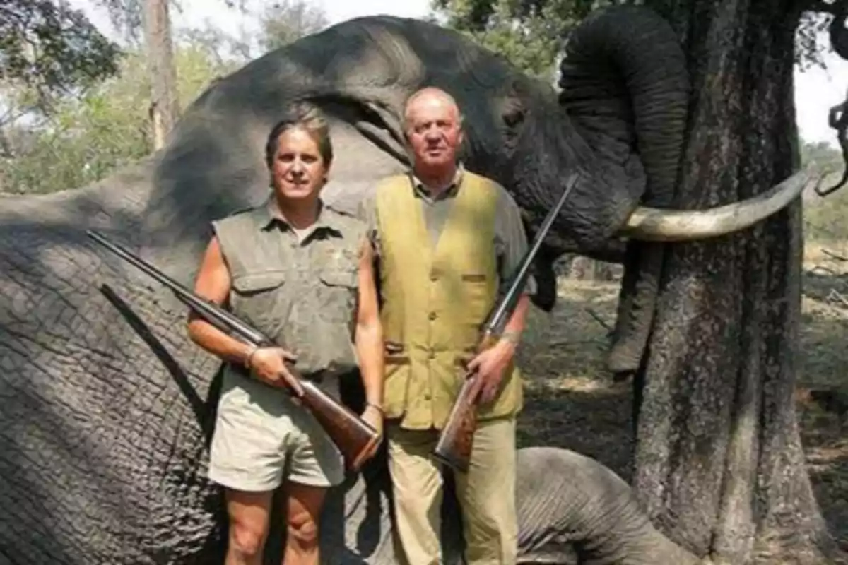 Dos hombres posan con rifles frente a un elefante muerto en un entorno de bosque.