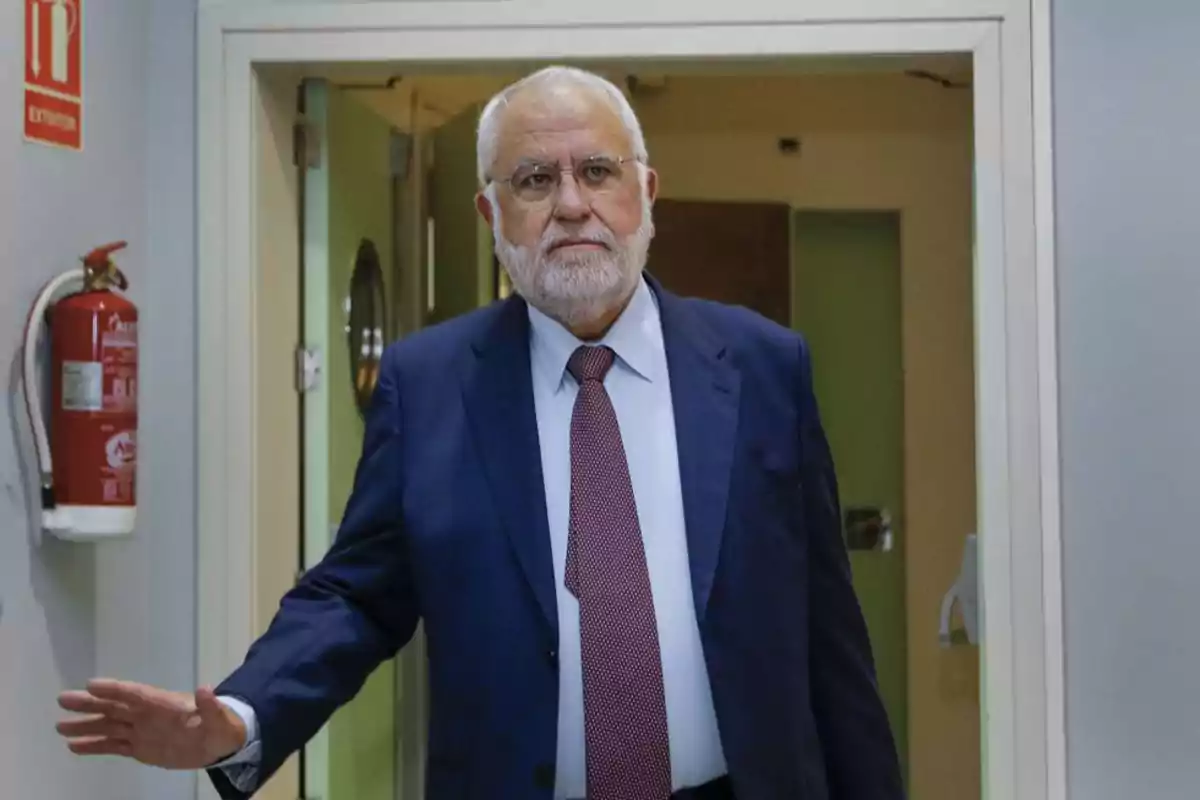 Hombre mayor con barba blanca y traje azul caminando por un pasillo con una puerta abierta detrás de él y un extintor de incendios en la pared. Hombre mayor con barba blanca y traje azul caminando por un pasillo con una puerta abierta detrás de él y un extintor de incendios en la pared.