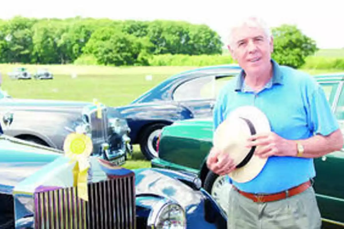 Un hombre de pie junto a un coche clásico sosteniendo un sombrero en un evento al aire libre.