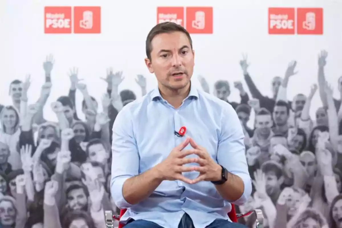 Un hombre sentado con camisa azul claro y micrófono en el pecho, frente a un fondo con logotipos de Madrid PSOE y una multitud en blanco y negro.