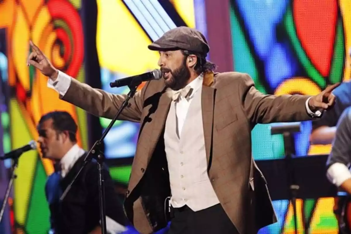 Un cantante con sombrero y chaqueta marrón actúa en un escenario colorido con un micrófono.