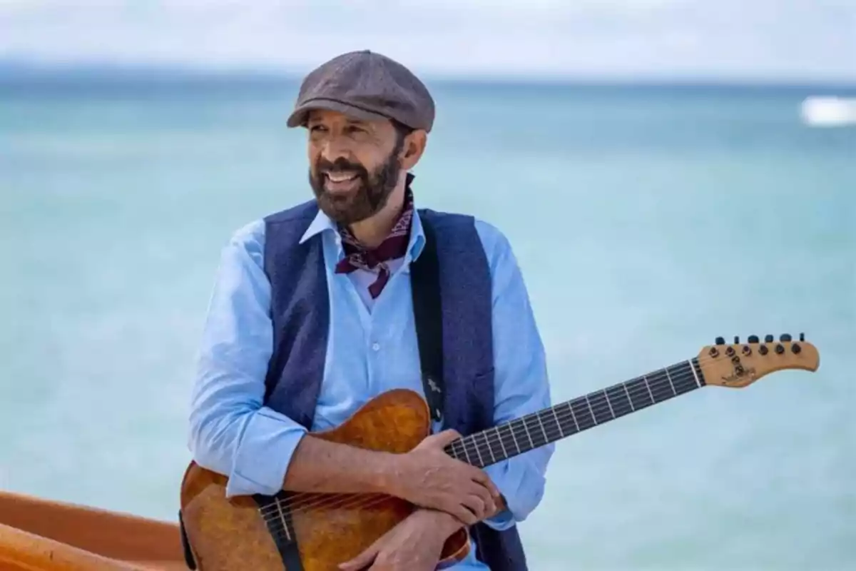 Un hombre con barba y sombrero sostiene una guitarra mientras sonríe en una playa con el mar de fondo.