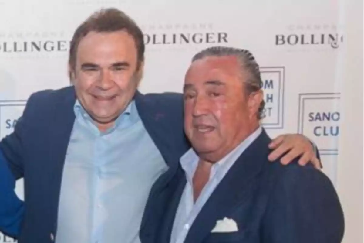 Dos hombres posando juntos en un evento con fondo de publicidad de Bollinger.