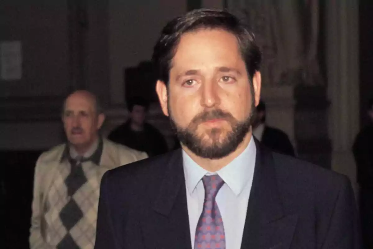 Un hombre con barba y traje oscuro, con una corbata de color rojo, está de pie en un entorno interior con otras personas en el fondo.