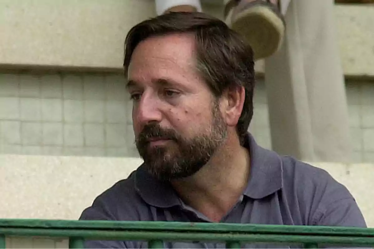 Hombre con barba y cabello oscuro sentado en una grada, vistiendo una camiseta polo gris.