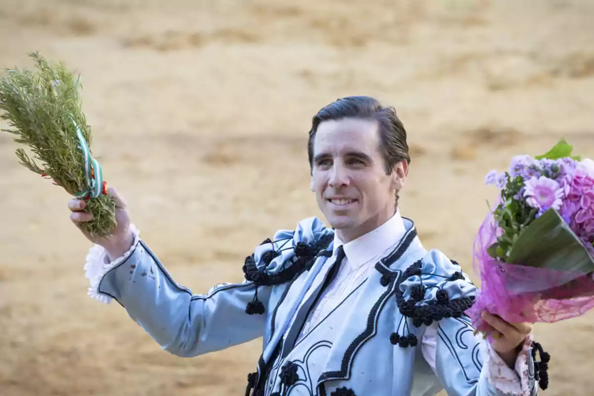 Un torero sonriente sostiene un ramo de flores y una rama de romero en una plaza de toros.