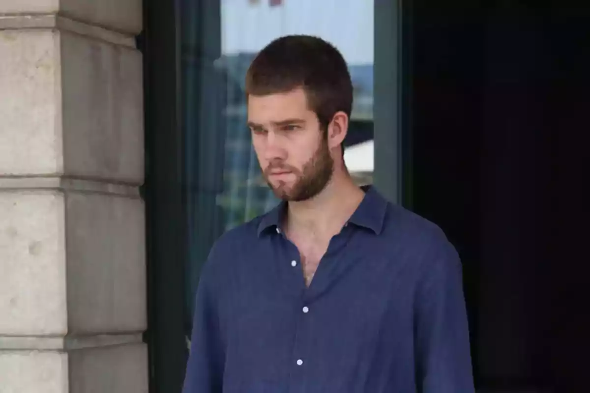 Un hombre con barba y cabello corto lleva una camisa azul mientras está de pie frente a un edificio.