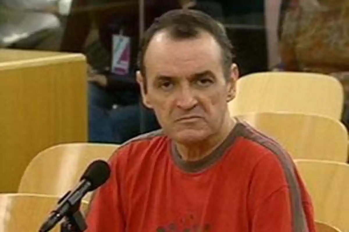 Un hombre con camiseta roja sentado frente a un micrófono en una sala con sillas de madera.