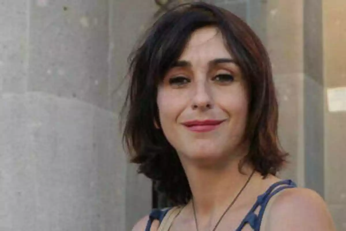 Una mujer con cabello oscuro y corto, sonriendo frente a un fondo de piedra.