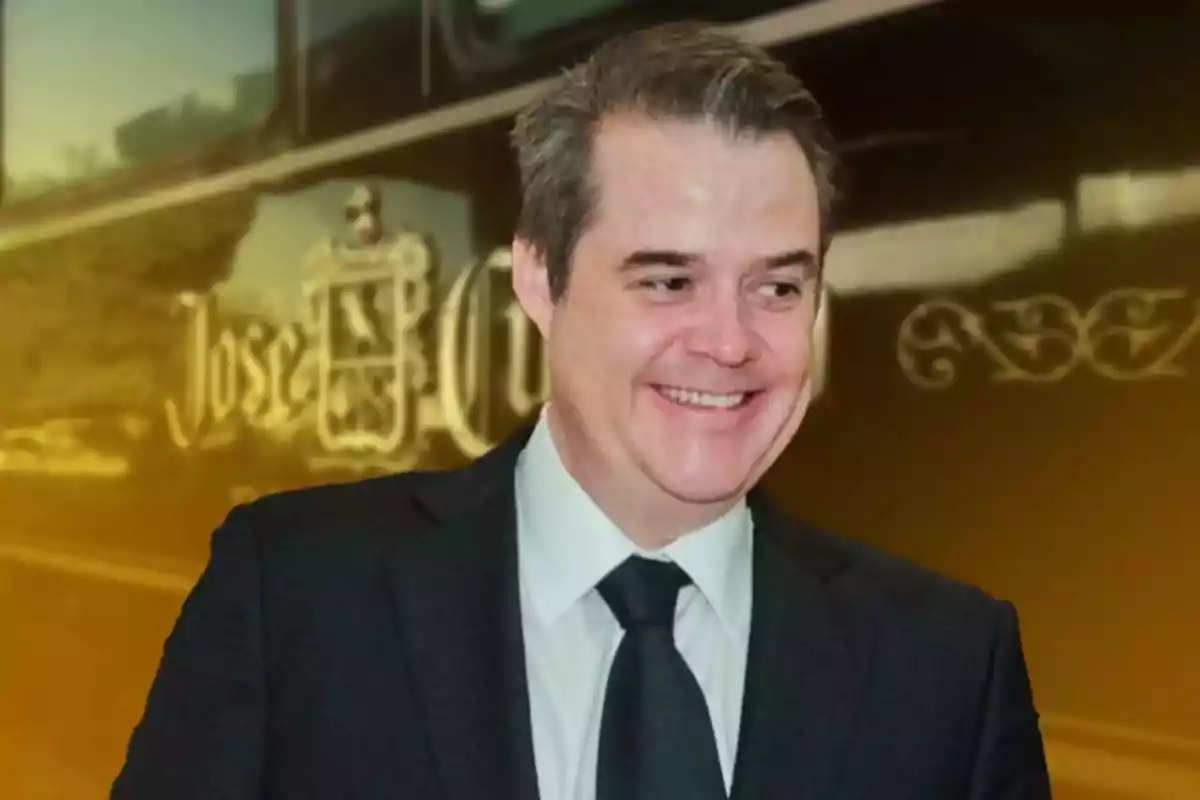 Un hombre sonriente con traje oscuro y corbata frente a un fondo con letras doradas. Un hombre sonriente con traje oscuro y corbata frente a un fondo con letras doradas.