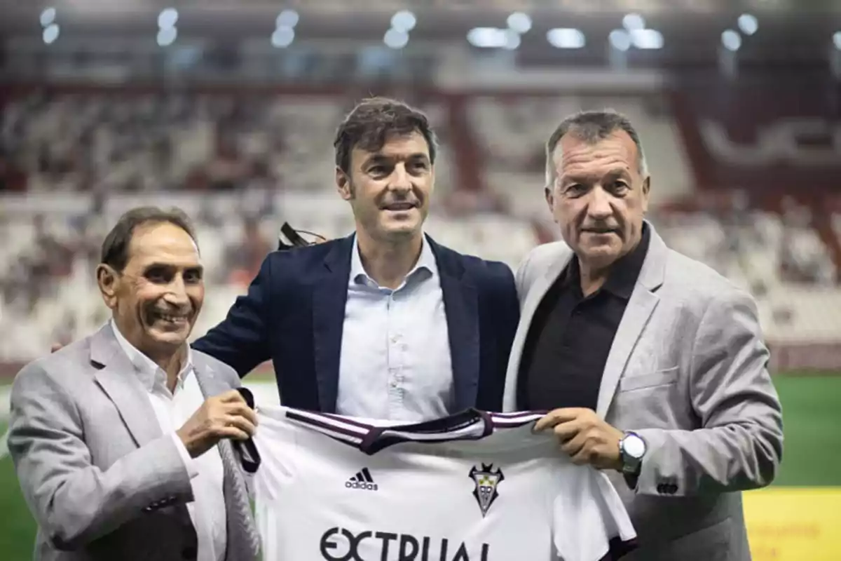 Tres hombres posan juntos en un estadio de fútbol, sosteniendo una camiseta del equipo Albacete Balompié.