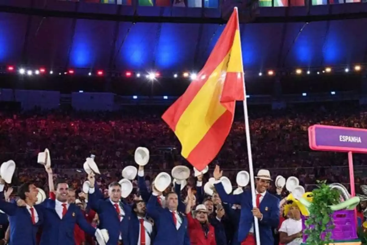 Un grupo de personas vestidas con trajes azules y corbatas rojas sostiene una bandera de España en un estadio iluminado.