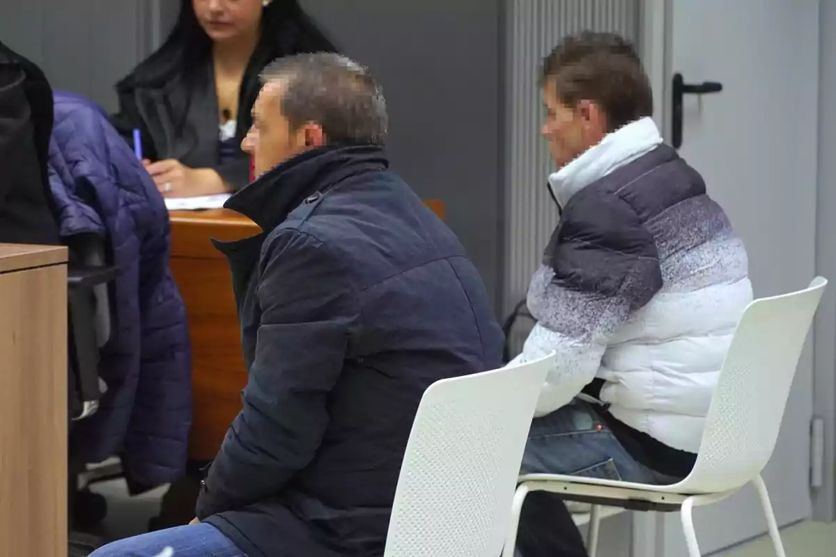 Dos personas sentadas en sillas blancas en una sala, con una mujer al fondo tomando notas.