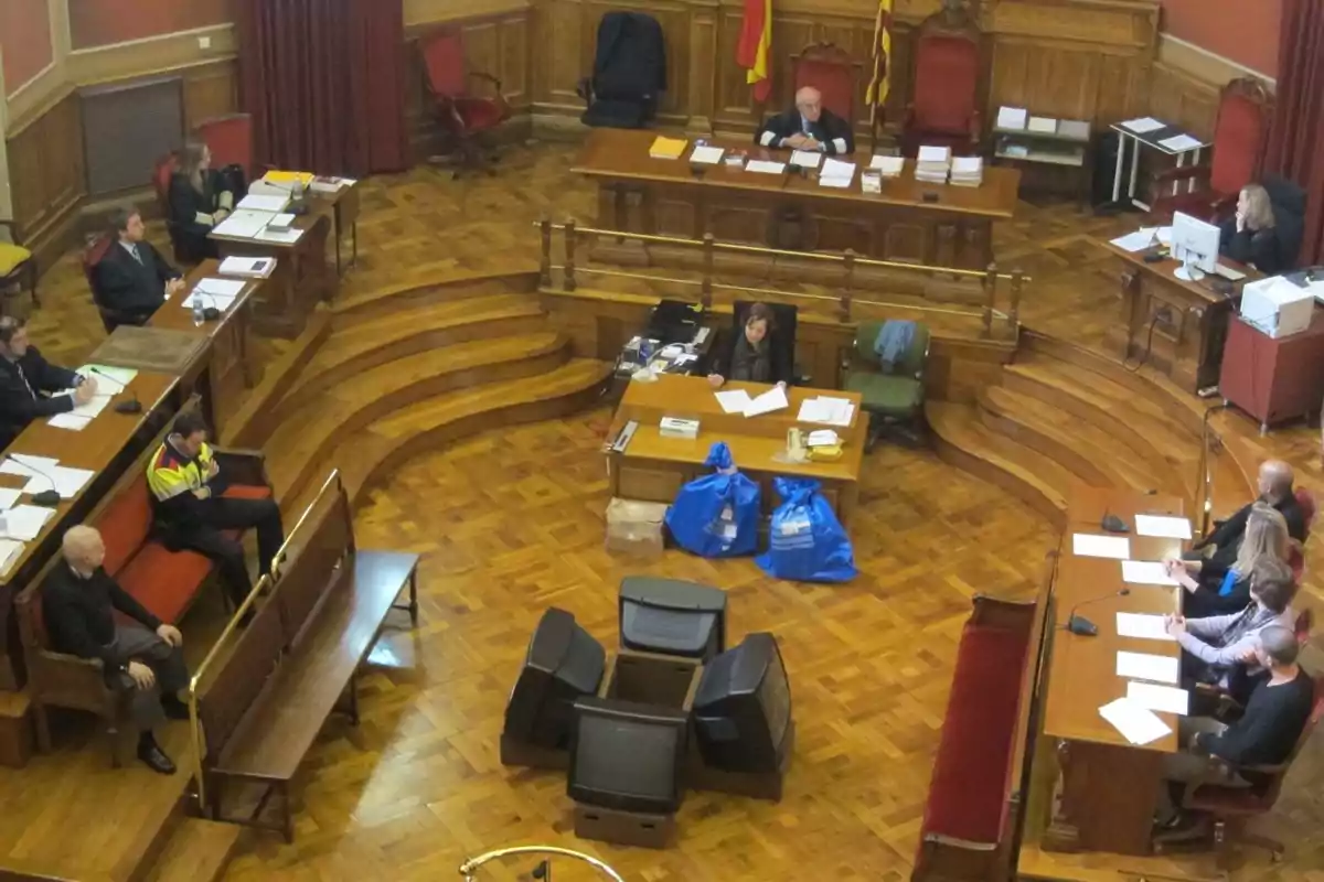 Una sala de tribunal con varias personas sentadas en escritorios de madera, un juez preside desde un estrado elevado, hay documentos y bolsas azules en el centro de la sala.