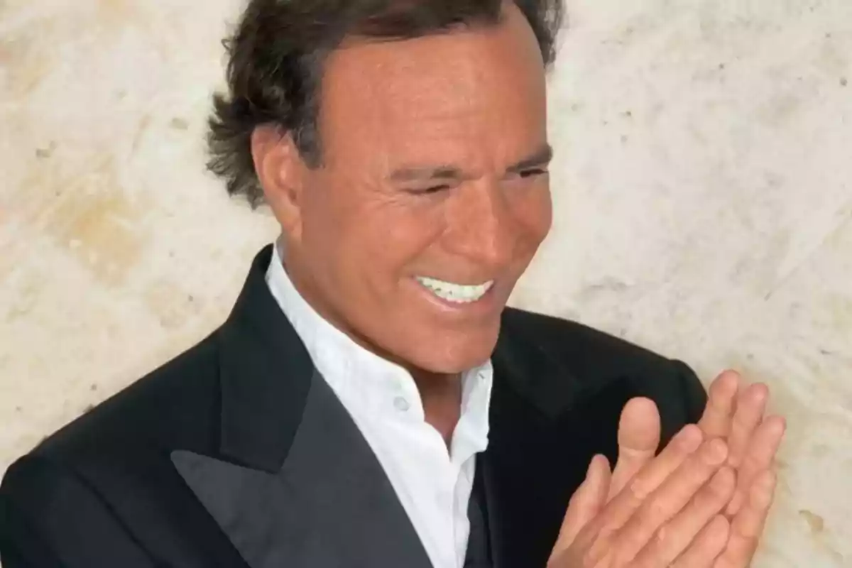 Hombre de cabello oscuro con traje negro y camisa blanca sonriendo y aplaudiendo frente a una pared clara