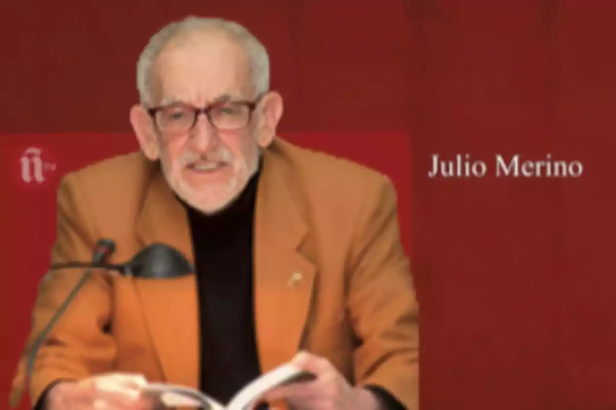 Un hombre mayor con gafas y barba blanca está sentado frente a un micrófono y sostiene un libro abierto, con un fondo rojo y texto que dice 