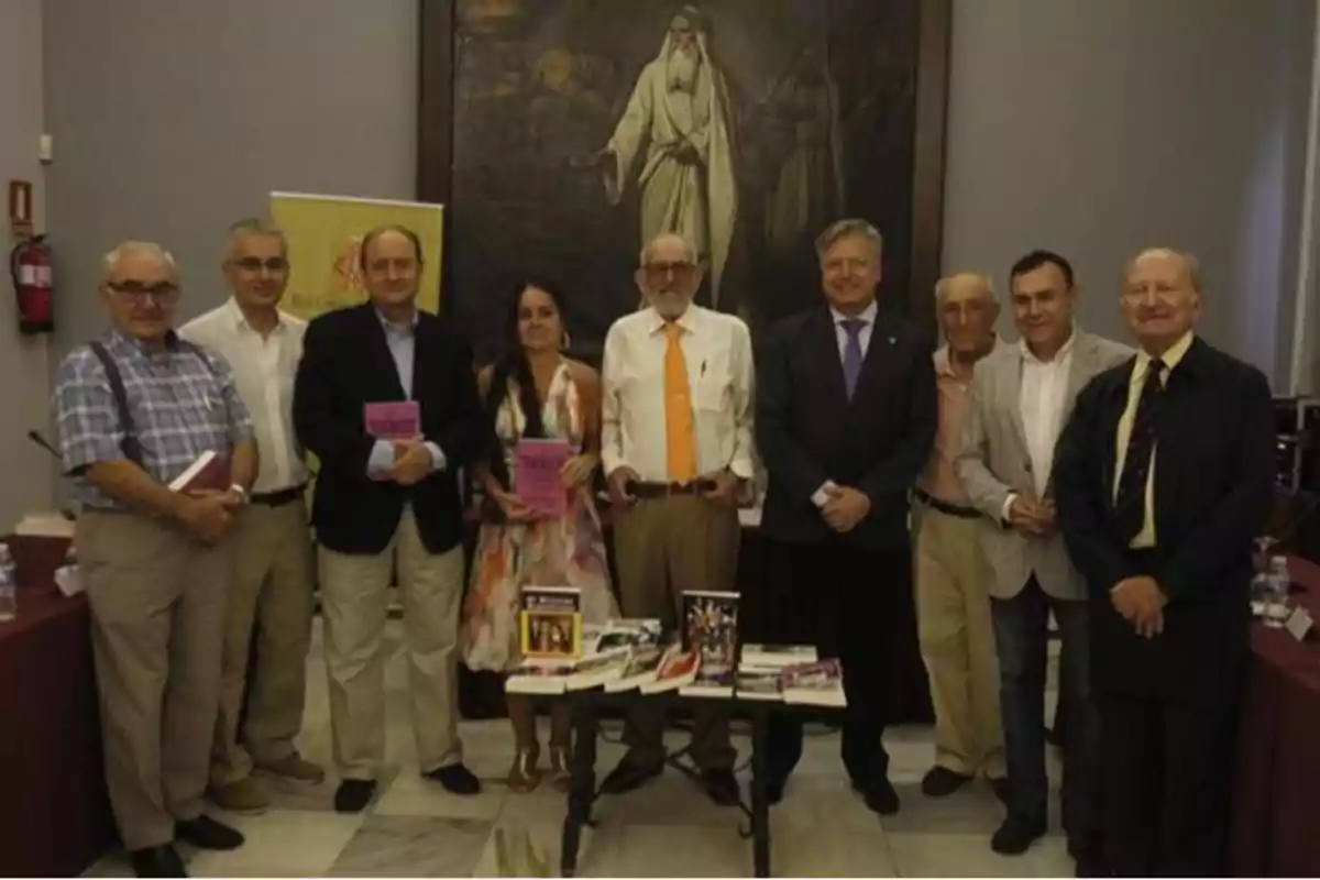 Un grupo de personas posando juntas en un evento, algunas sosteniendo libros, con una pintura grande en la pared detrás de ellas.