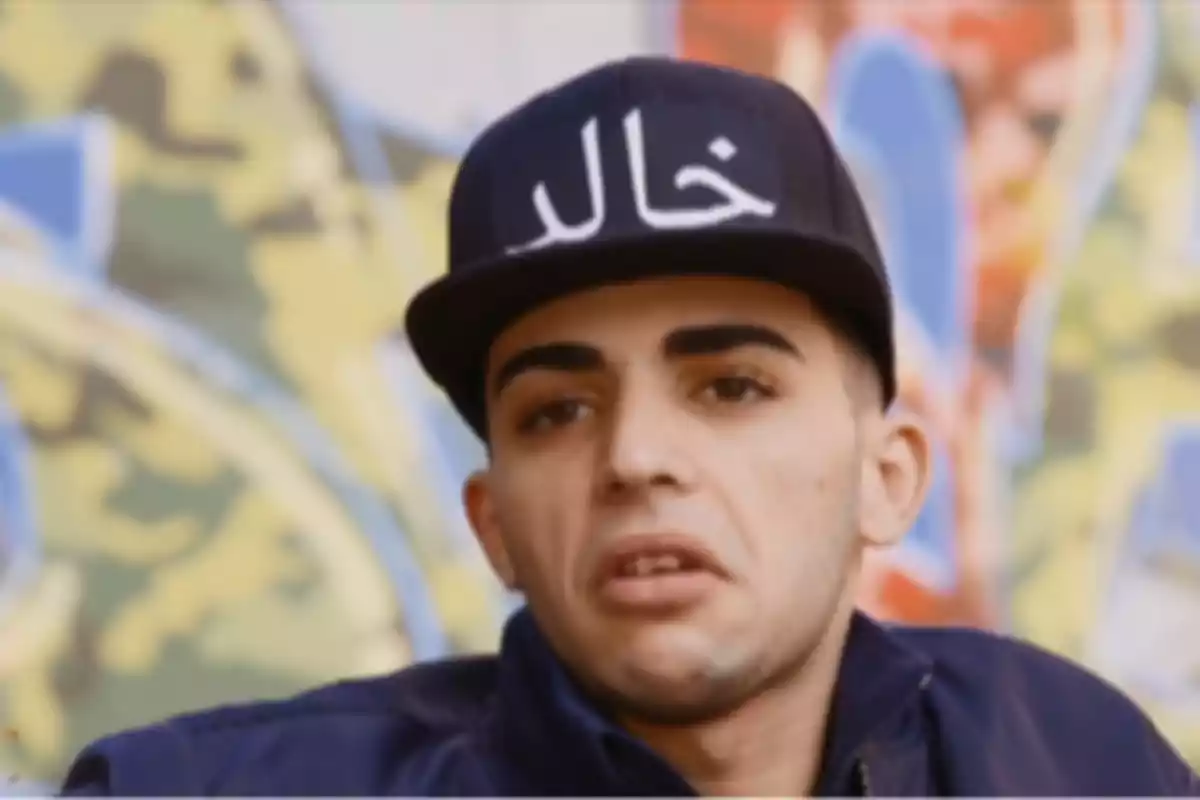 Un hombre con gorra negra con escritura blanca y fondo de graffiti colorido.