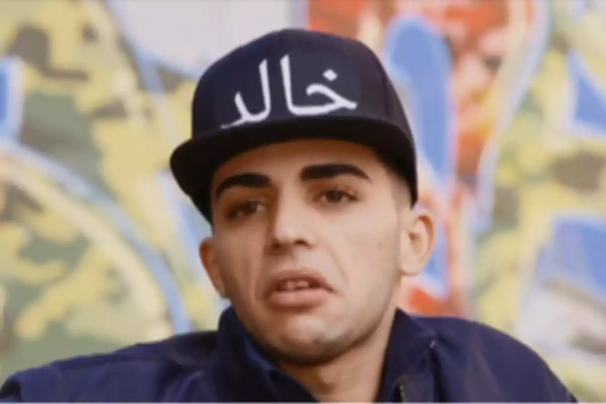 Un hombre con gorra negra con letras blancas y fondo de graffiti colorido.