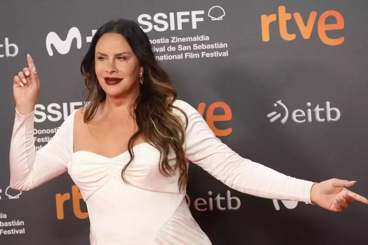 Una mujer con vestido blanco posando en una alfombra roja frente a un fondo con logotipos de un festival de cine.