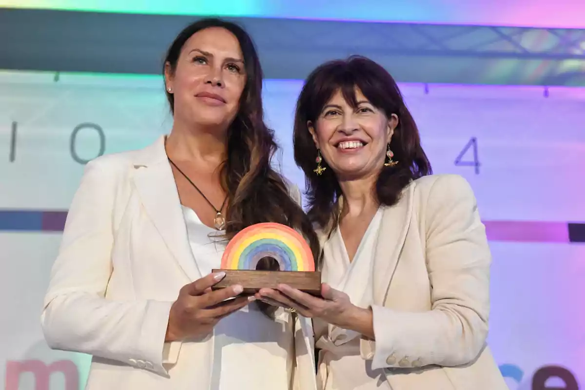 Dos mujeres sonrientes sostienen un trofeo con forma de arcoíris en un evento.