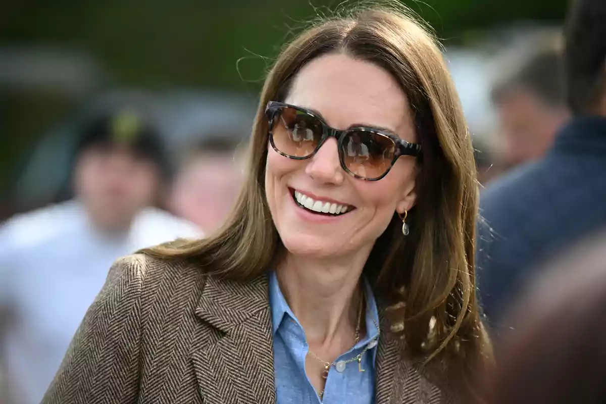 Kate Middleton smiling with sunglasses and long hair, wearing a brown blazer and a blue jersey, in an outdoor setting. Kate Middleton smiling with sunglasses and long hair, wearing a brown blazer and a blue jersey, in an outdoor setting.