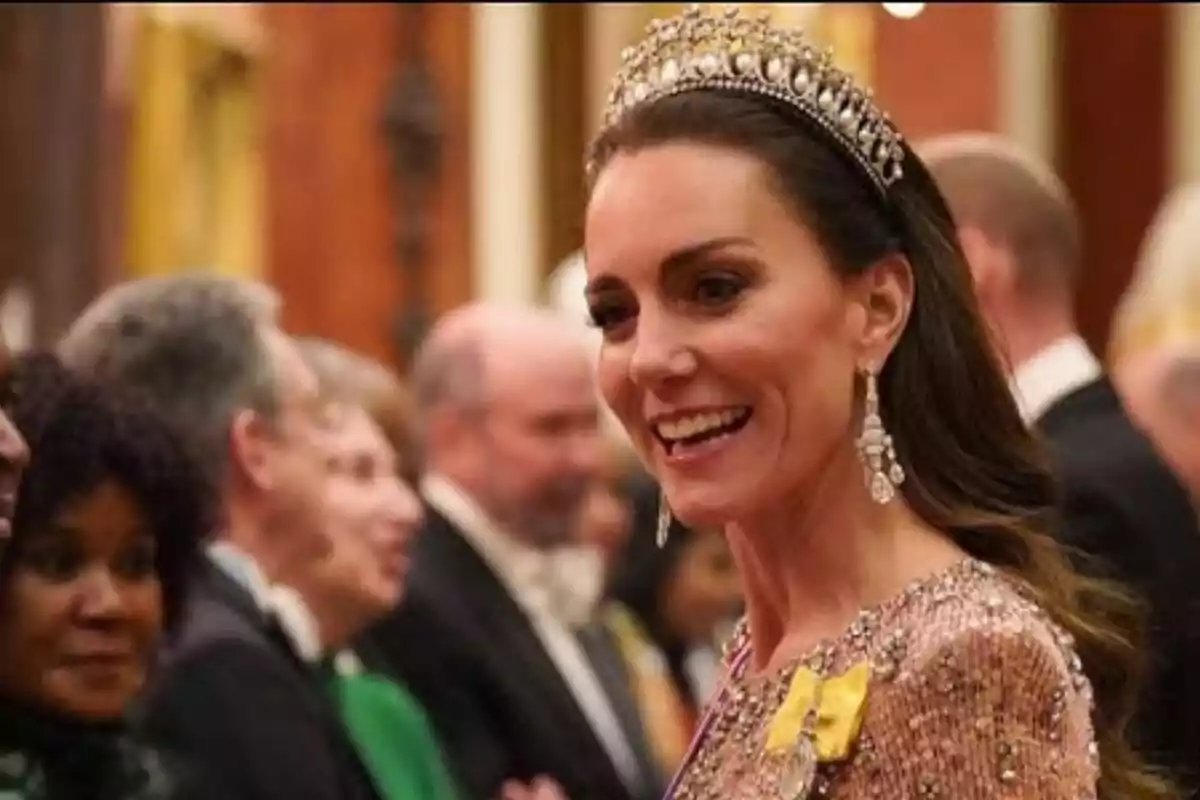 Kate Middleton con tiara y vestido elegante sonríe en un evento formal rodeada de otras personas.